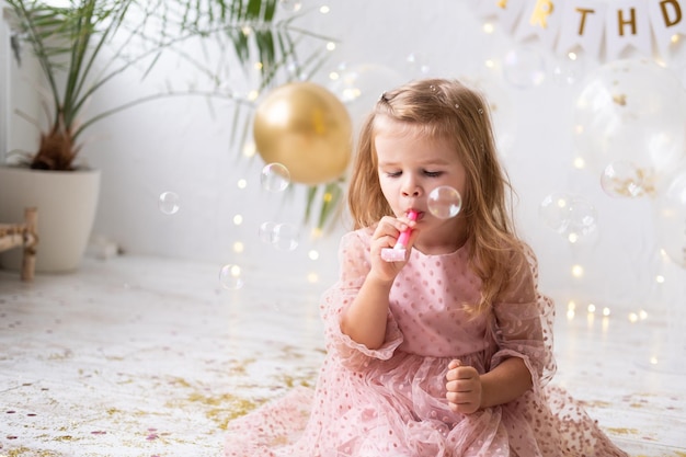 Bambina felice con i capelli lunghi in abito rosa che soffia in un fischietto e festeggia il suo compleanno