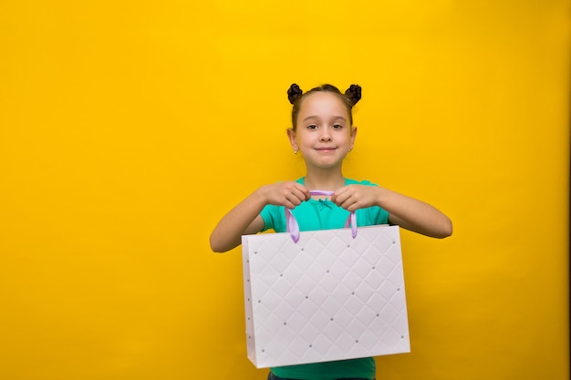 Bambina felice con le code divertenti che stanno isolate sopra i sacchetti della spesa gialli della tenuta della parete. Guardando la fotocamera