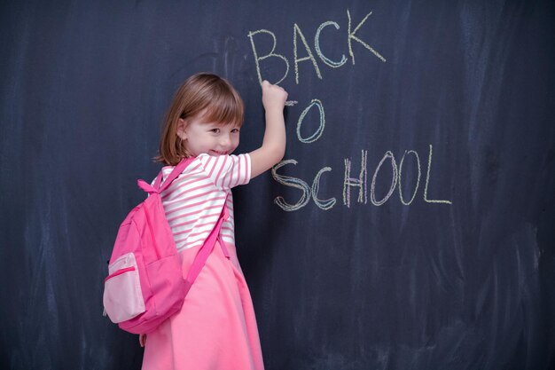 Bambina felice della scuola con lo zaino che scrive di nuovo a scuola sulla lavagna nera