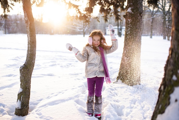 Bambina felice di 5-6 anni che gioca con la neve sopra la luce del sole nel parco innevato all'aperto. Il bambino allegro indossa una giacca invernale e una sciarpa. Infanzia.