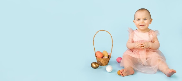 Bambina felice di dieci mesi che indossa la fascia delle orecchie del coniglietto che si siede sullo sfondo azzurro dello studio e gioca con le uova di Pasqua colorate Biglietto di auguri di Pasqua per bambini piccoli Coniglietto di pasqua del bambino