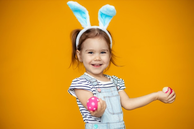 Bambina felice divertente con uova di Pasqua e orecchie da coniglio su sfondo giallo