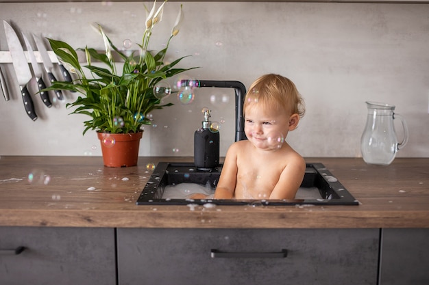 Bambina felice e carina che gioca con l'acqua e la schiuma in un lavello da cucina a casa