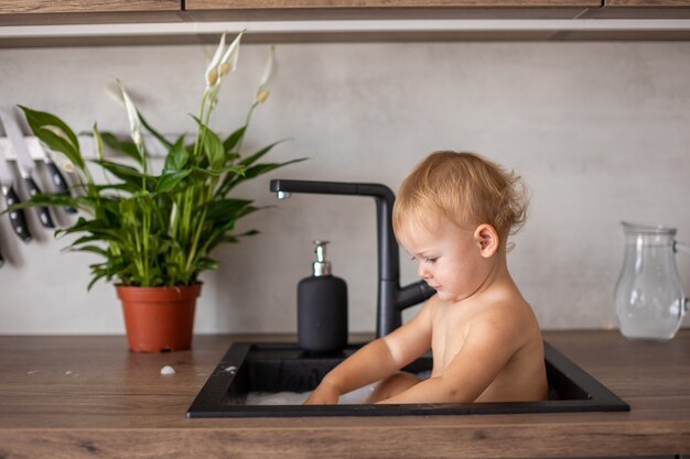 Bambina felice e carina che gioca con l'acqua e la schiuma in un lavello da cucina a casa