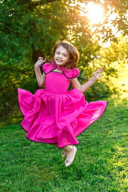 Bambina felice in abito rosa che sorride e salta sull'erba in estate al tramonto