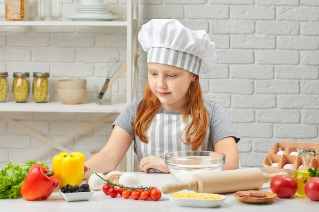 Bambina felice in cappello da chef e un grembiule che cucina la pizza