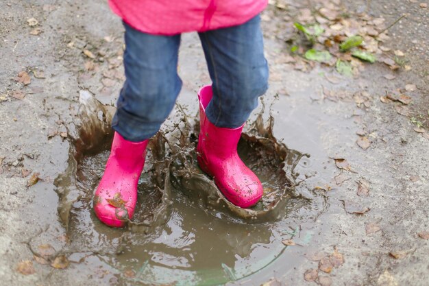 Bambina felice in giacca impermeabile rosa stivali di gomma salta allegramente attraverso le pozzanghere sulla strada in caso di pioggia Primavera autunno Divertimento per bambini all'aria aperta dopo la pioggia Ricreazione all'aperto