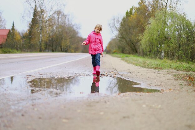 Bambina felice in giacca impermeabile rosa stivali di gomma salta allegramente attraverso le pozzanghere sulla strada in caso di pioggia Primavera autunno Divertimento per bambini all'aria aperta dopo la pioggia Ricreazione all'aperto