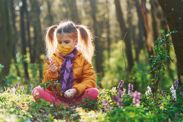 Bambina felice in maschera protettiva seduta nella foresta primaverile durante il giorno