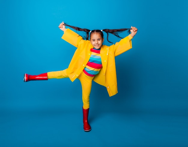 Bambina felice in un impermeabile giallo e stivali rossi su sfondo blu.