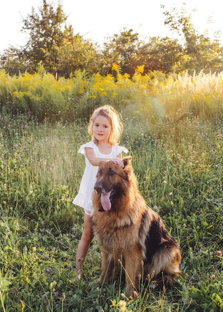 Bambina felice in vestito bianco che accarezza il grande cane che sta sull'erba verde in primavera. Pastore tedesco