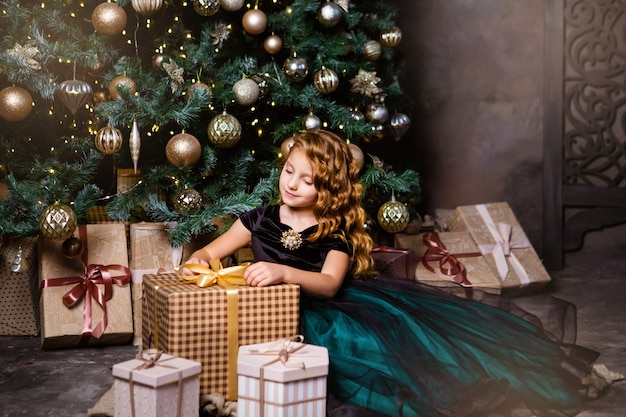 Bambina felice in vestito operato che si siede vicino all'albero di Natale con la scatola attuale. Tempo di Natale e concetto di Capodanno. Colori verdi e dorati delle decorazioni.