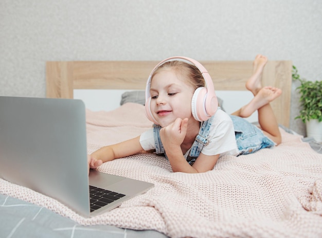 Bambina felice sorridente che usa il computer portatile in cuffie wireless a letto a casa.