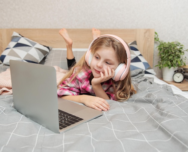 Bambina felice sorridente che usa il computer portatile in cuffie wireless a letto a casa