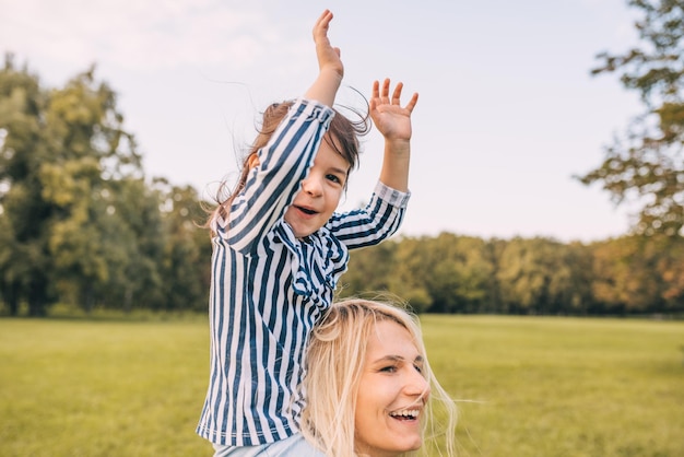 Bambina felice su un piggy back con le mani in alto giro con sua madre sorridente sullo sfondo della natura Donna amorevole e la sua bambina che giocano nel parco Mamma e bambino si divertono fuori Maternità