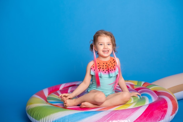 Bambina felice sveglia in costume da bagno sorridente seduta sul materasso gonfiabile colorato.