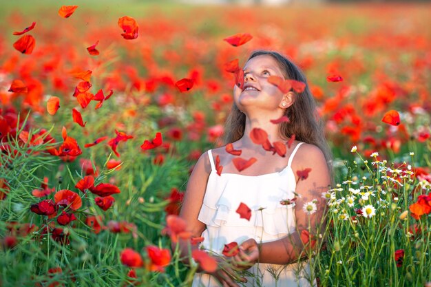 Bambina graziosa che indossa un abito bianco in un campo di papaveri in fiore estivo che gioca sotto il sole della sera