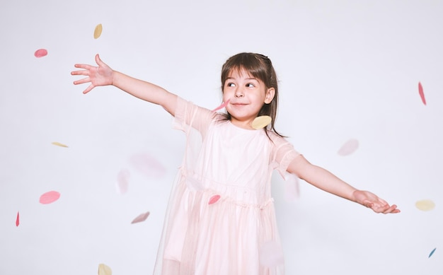 Bambina graziosa che indossa un abito rosa in tulle con corona di principessa sulla testa isolata su sfondo bianco alza le mani goditi la sorpresa dei coriandoli Ragazza sorridente carina che celebra la sua festa di compleanno divertendosi