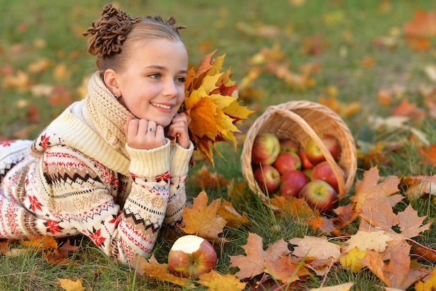 Bambina graziosa che si trova sull'erba verde vicino al cesto di vimini pieno di mele