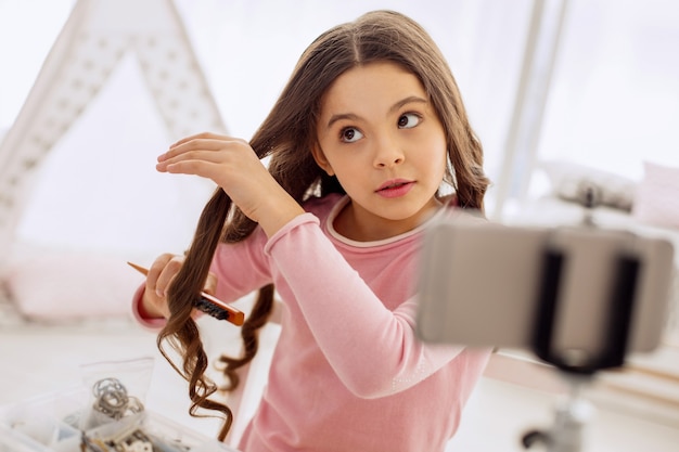 Bambina graziosa che spazzola i suoi capelli davanti alla macchina fotografica