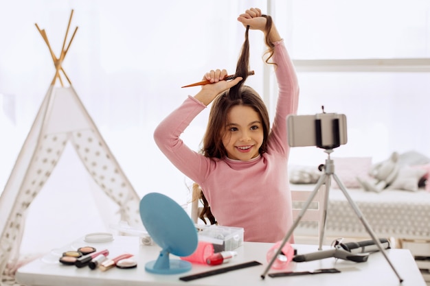 Bambina graziosa che spazzola i suoi capelli davanti alla macchina fotografica