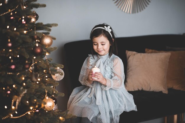 Bambina graziosa che tiene una candela accesa seduta su un albero decorato di Natale
