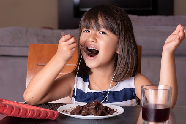 Bambina graziosa felice che guarda tablet e mangia la torta al cioccolato