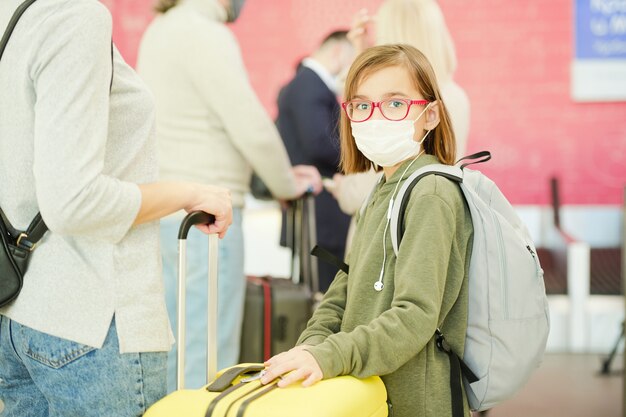 Bambina in abbigliamento casual, occhiali e maschera protettiva in piedi davanti alla telecamera contro i viaggiatori che aspettano il loro turno per il check-up in aeroporto