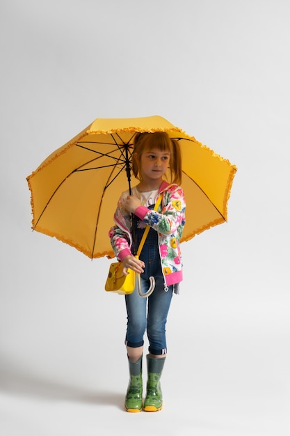 Bambina in abiti autunnali casual e stivali di gomma finge di nascondersi dalla pioggia sotto un ombrello giallo su sfondo bianco