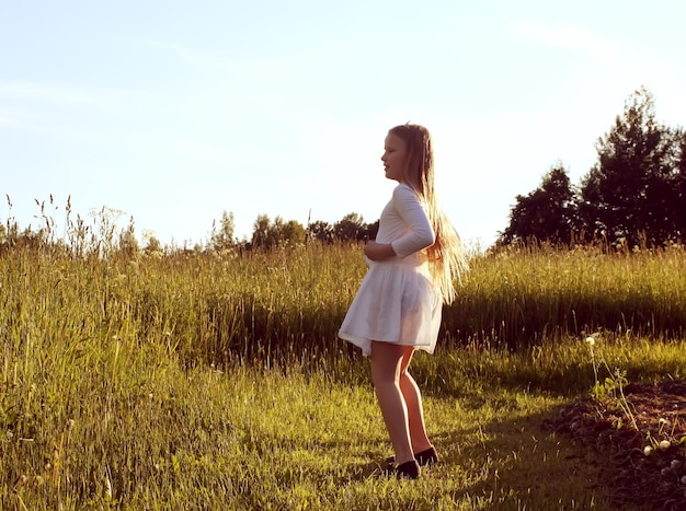 Bambina in abito bianco che cammina all'aperto sul campo estivo verde in campagna