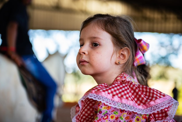 Bambina in abito scozzese rosso e fiocco di nastro tra i capelli con cavallo sfocato sullo sfondo.