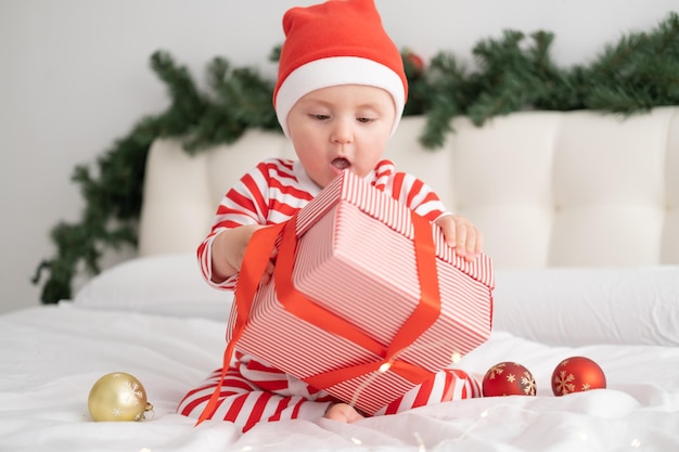 Bambina in body a righe e cappello di Babbo Natale che gioca con una confezione regalo sulla camera da letto decorata di Natale