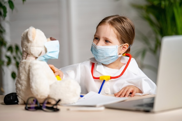 Bambina in camice bianco e maschera, usando lo stetoscopio, ascolta orsacchiotti.
