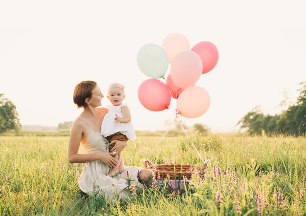 Bambina in cesto di vimini con palloncini alla luce del sole al tramonto Madre e bambino all'aperto Primo compleanno La famiglia festeggia un bambino di un anno sulla natura Foto di sogni sani vacanze vacanze