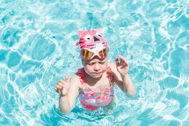 Bambina in coppa rosa da bagno e occhiali che nuotano nella piscina all'aperto in estate.