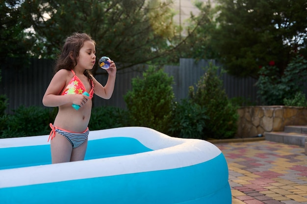 Bambina in costume da bagno soffia bolle di sapone mentre prende il sole in una piscina gonfiabile nel giardino sul retro della casa