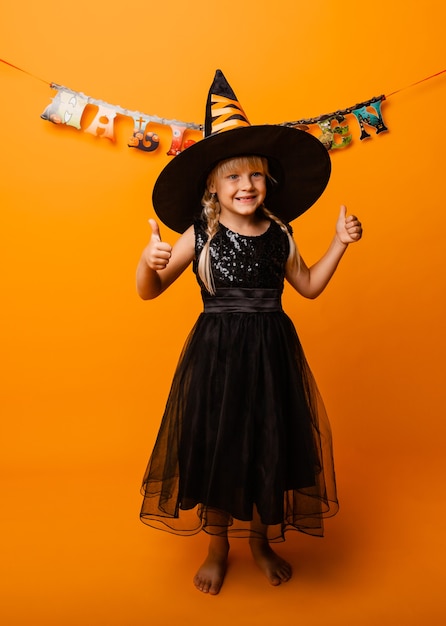 Bambina in costume di halloween nero che ride e guarda la telecamera, salta e si diverte, isolata su sfondo giallo. Halloween