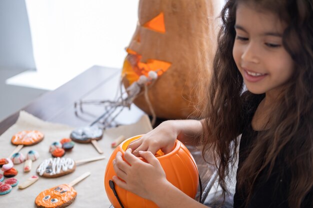 Bambina in costume di strega che tiene la presa della zucca con le caramelle, divertendosi in cucina, celebrando Halloween. messa a fuoco selettiva.