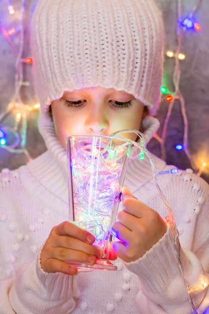 Bambina in maglione bianco e cappello bevande da un vetro multicolore luci di una ghirlanda di Natale. Concetto creativo di umore di natale.