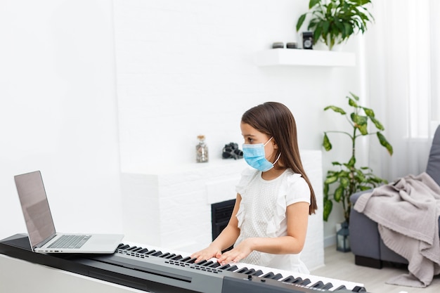 Bambina in maschera protettiva che impara il pianoforte durante la quarantena. Concetto di coronavirus.