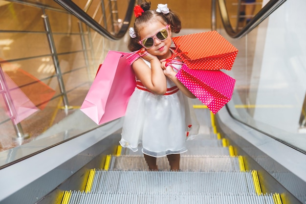 Bambina in occhiali da sole sulla scala mobile nel centro commerciale con gli acquisti