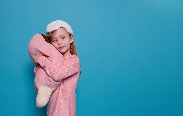 Bambina in pigiama e maschera per dormire abbraccia il giocattolo su uno spazio di copia sfondo blu
