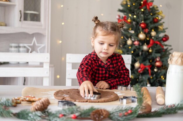 Bambina in pigiama rosso che cucina pan di zenzero festivo in cucina decorata natalizia