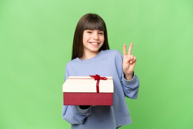 Bambina in possesso di un regalo su sfondo chroma key isolato sorridente e mostrando segno di vittoria