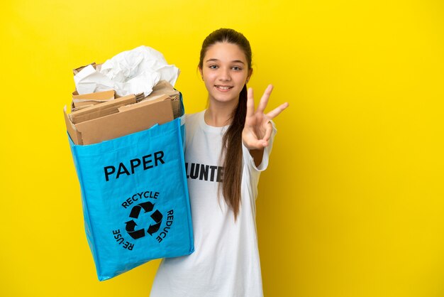 Bambina in possesso di un sacchetto di riciclaggio pieno di carta da riciclare su sfondo giallo isolato felice e contando tre con le dita