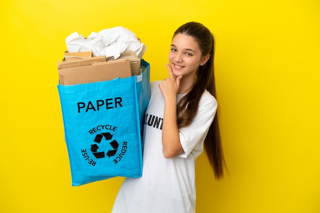 Bambina in possesso di un sacchetto di riciclaggio pieno di carta da riciclare su sfondo giallo isolato felice e sorridente