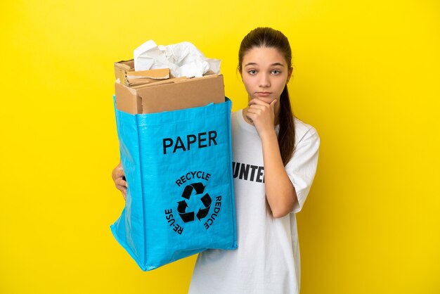 Bambina in possesso di un sacchetto di riciclaggio pieno di carta da riciclare su sfondo giallo isolato pensando