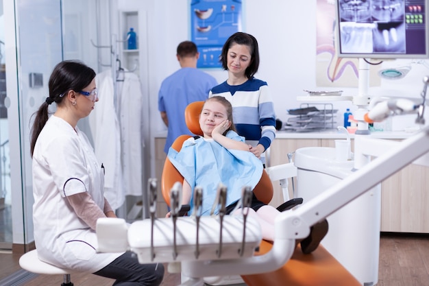 Bambina in studio dentistico pediatrico toccando il viso con espressione dolorosa a causa di problemi di salute dei denti. Bambino con sua madre durante il controllo dei denti con stomatolog seduto su una sedia.