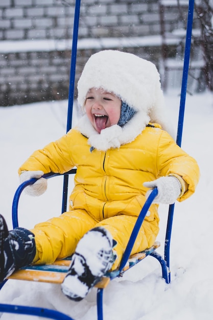 bambina in tuta da neve gialla che attacca fuori la lingua, seduta in slitta in una giornata invernale nevosa