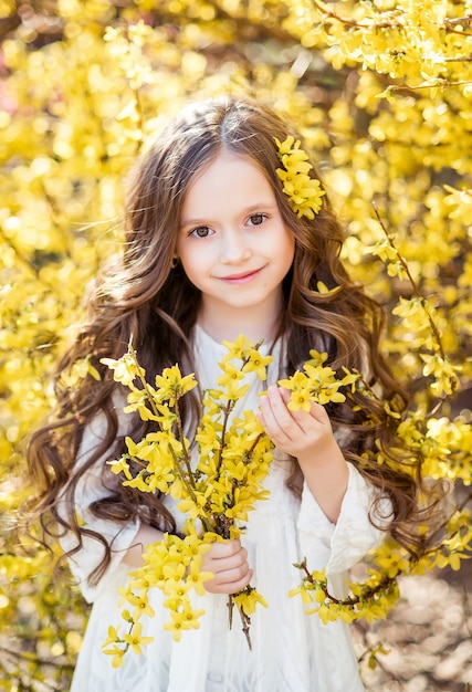 Bambina in un abito bianco con fiori gialli nelle sue mani. Un bambino su uno sfondo di fiori gialli. Ritratto di primavera di un bambino ..